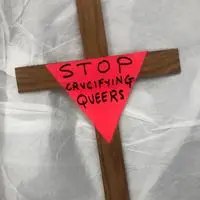 A wooden cross with a pink triangle on that reads "STOP CRUCIFYING QUEERS"