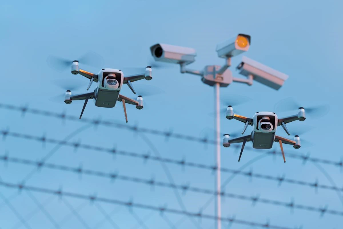 Two small drones in front of barbed wire. Three CCTV cameras are out of focus in the background.