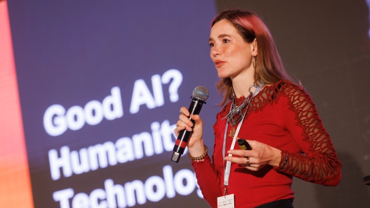 Photograph of Eleanor Drage holding a microphone in her right hand delivering a speech while standing in front of a projected image thta reads Good AI? Humanity, Technology, Justice.