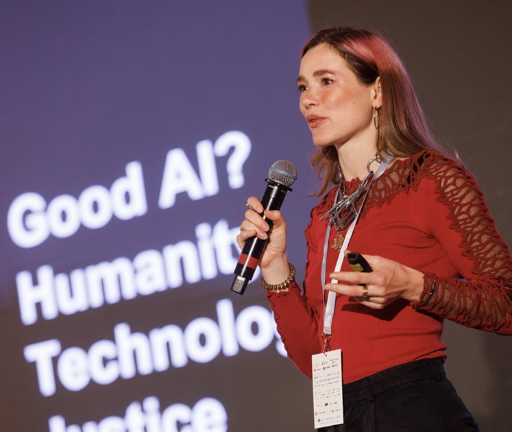 Photograph of Eleanor Drage holding a microphone in her right hand delivering a speech while standing in front of a projected image thta reads Good AI? Humanity, Technology, Justice.