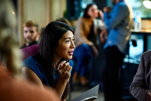 Woman in a busy office with a notebook in hand speaking to someone out of shot