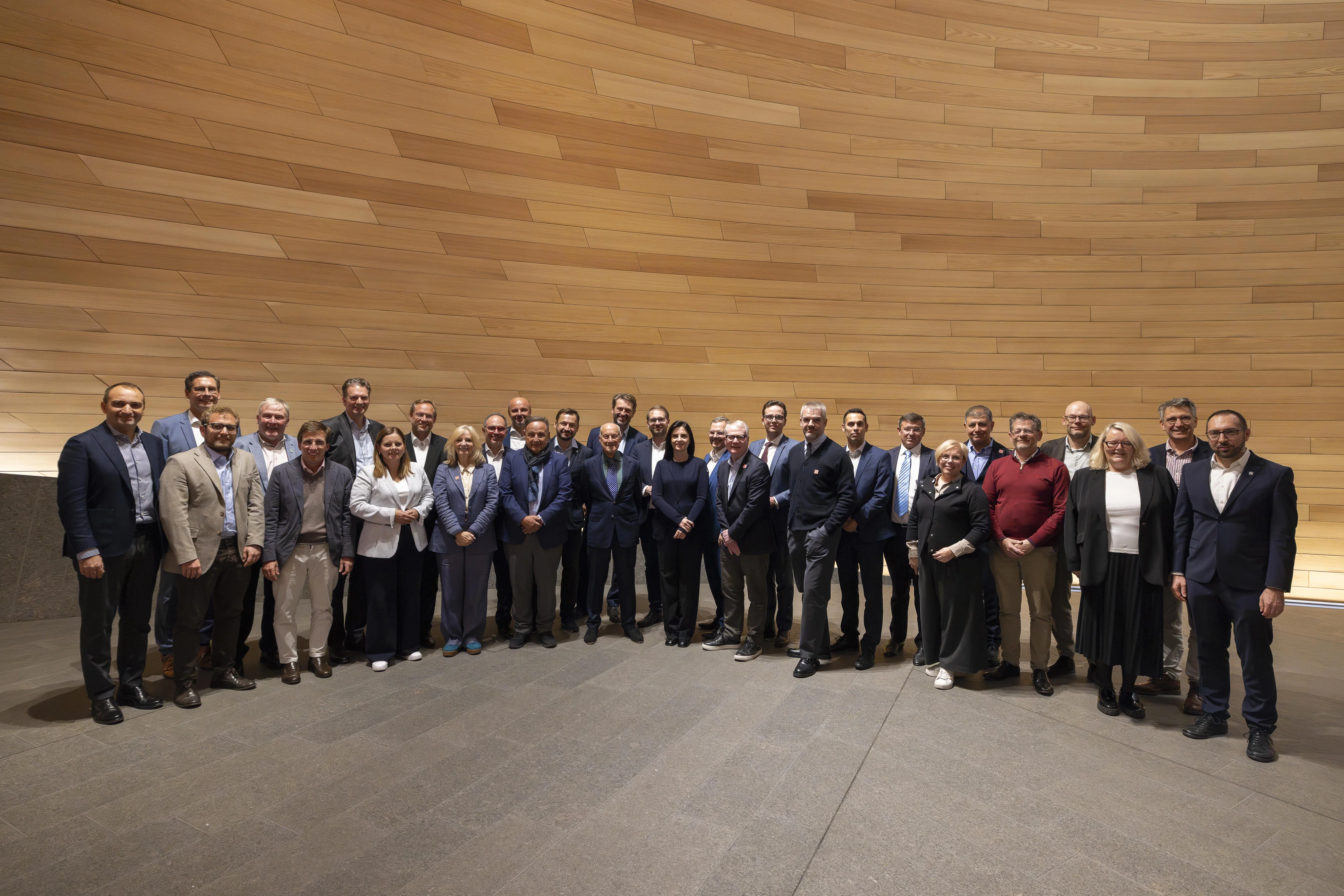 Group of mayors and city leaders from across Europe gather in a semi circle in front of a modern wood-panneled backdrop indoors.