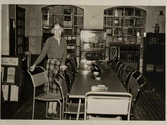 A person stood at a long desk pulling one of the chairs away from the table.