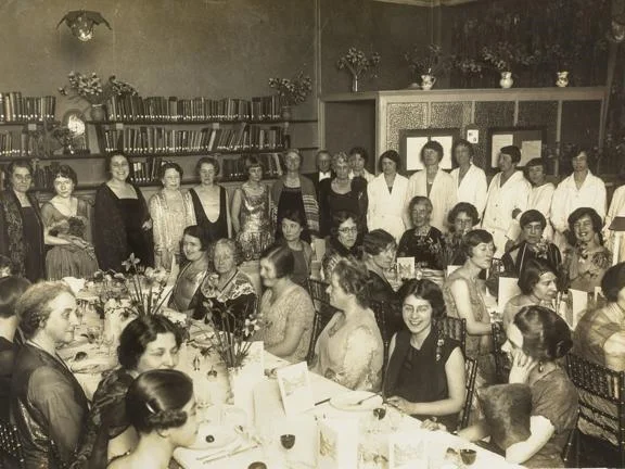 A large group photo, some people stood and others sat at dressed tables.