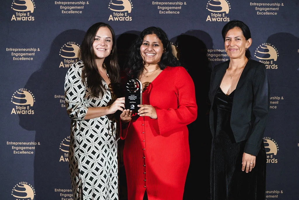 Three women smile at the camera holding an award trophy from the Triple E awards. There is a branded screen behind them with the Triple E name and logo.
