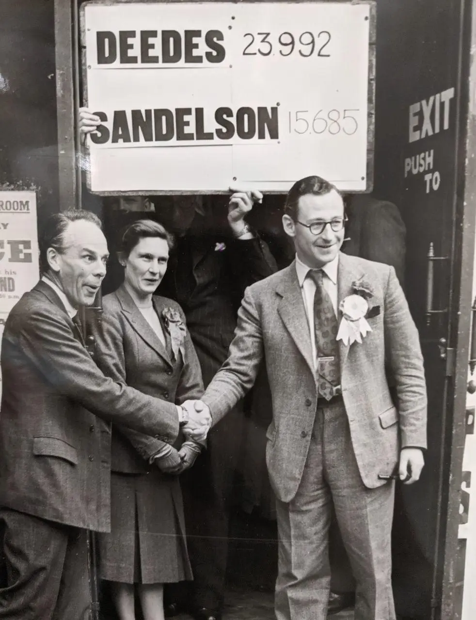 Two politicians shaking hands below a vote count.
