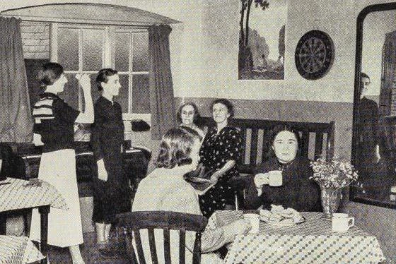 A club room with people sat at tables and others playing darts.