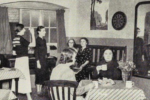 A club room with people sat at tables and others playing darts.