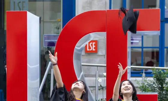 Two female graduates throwing their mortar boards in the air in front of large red L S E letters