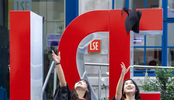 Two female graduates throwing their mortar boards in the air in front of large red L S E letters
