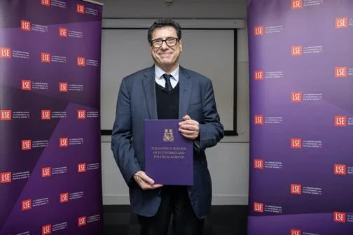 Philippe Aghion, standing between two LSE banners, holding a gift book, given in celebration of receiving Nobel prize