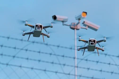 Two small drones in front of barbed wire. Three CCTV cameras are out of focus in the background.