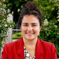 Rabia is smiling and facing the camera. She is wearing a red blazer with gold jewellery.