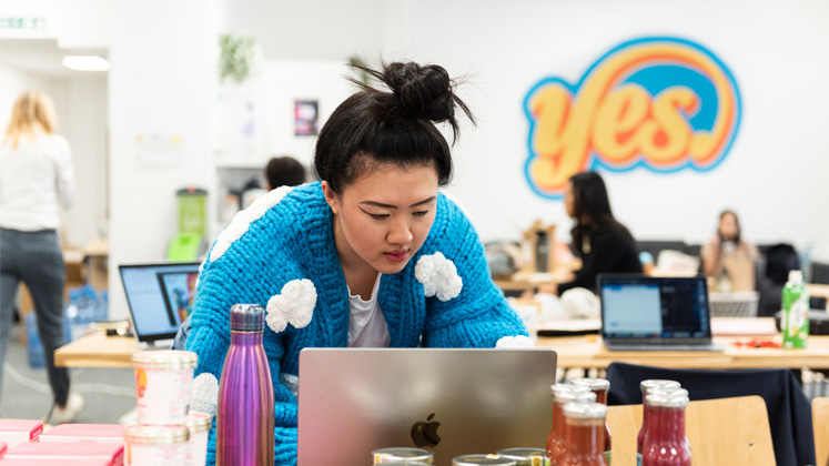 A young woman wearing a bright blue cardigan works on a laptop in a busy office with a yellow painting on the wall that says ''yes'.