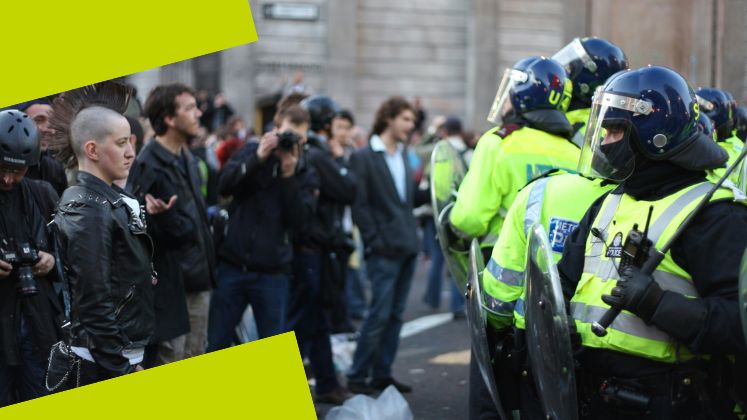 A group of people in black clothing stand in front of a row of police in riot wear