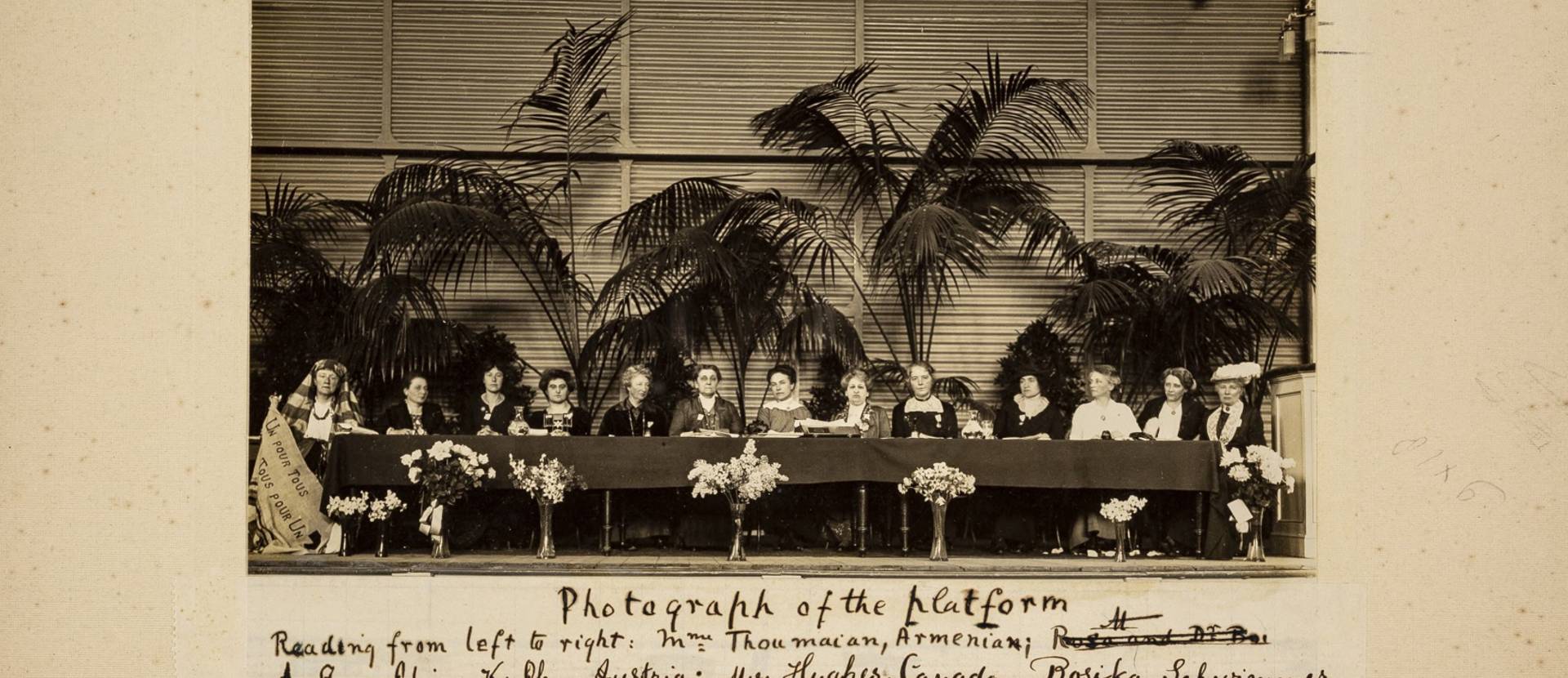 A long desk on a platform with a line of women sat at it. Palm trees in the background