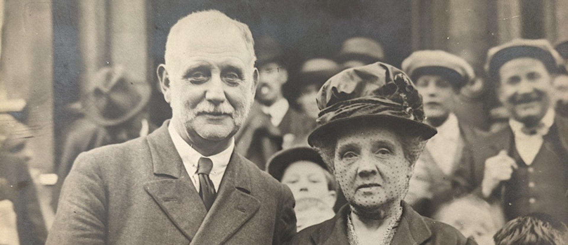 George Lansbury stood with his wife for a photograph. Behind them is a crowd of people.