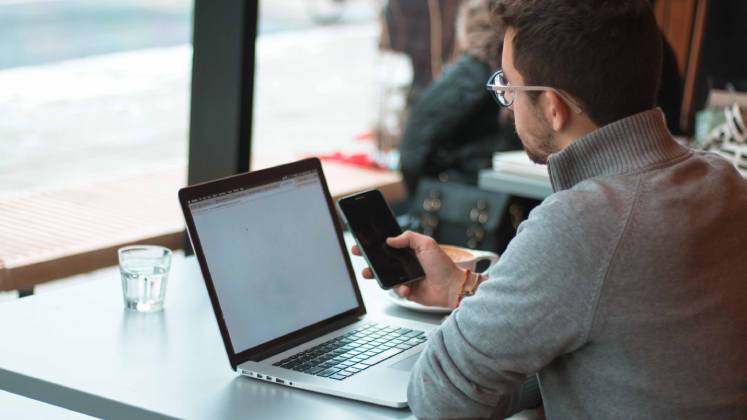 Man sits in a cafe with his laptop open. He holds his phone in his hand.